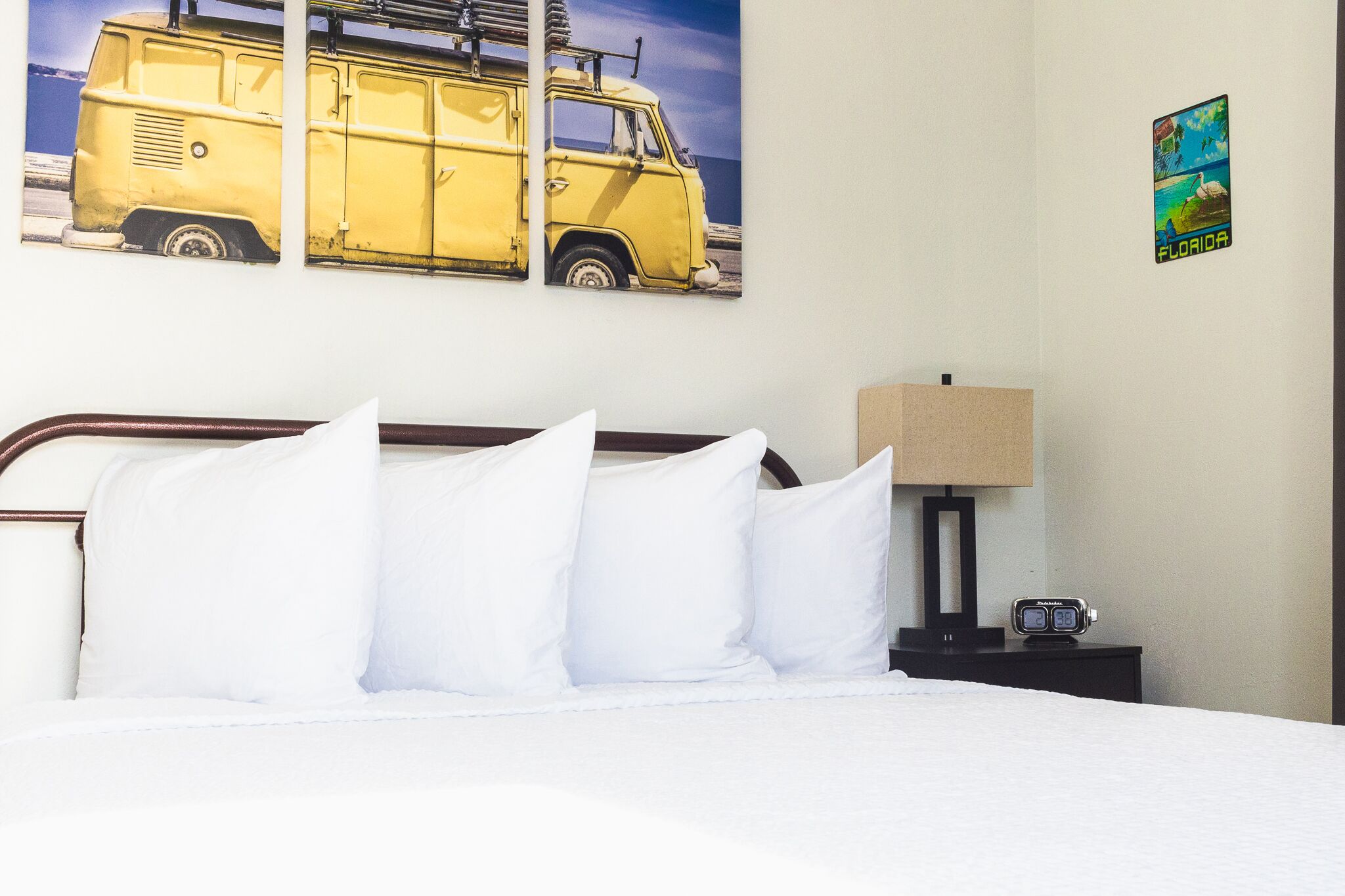 A hotel room with a bed, pillows, a nightstand with a lamp, a clock, and two wall art pieces depicting a yellow van and a Florida landscape.