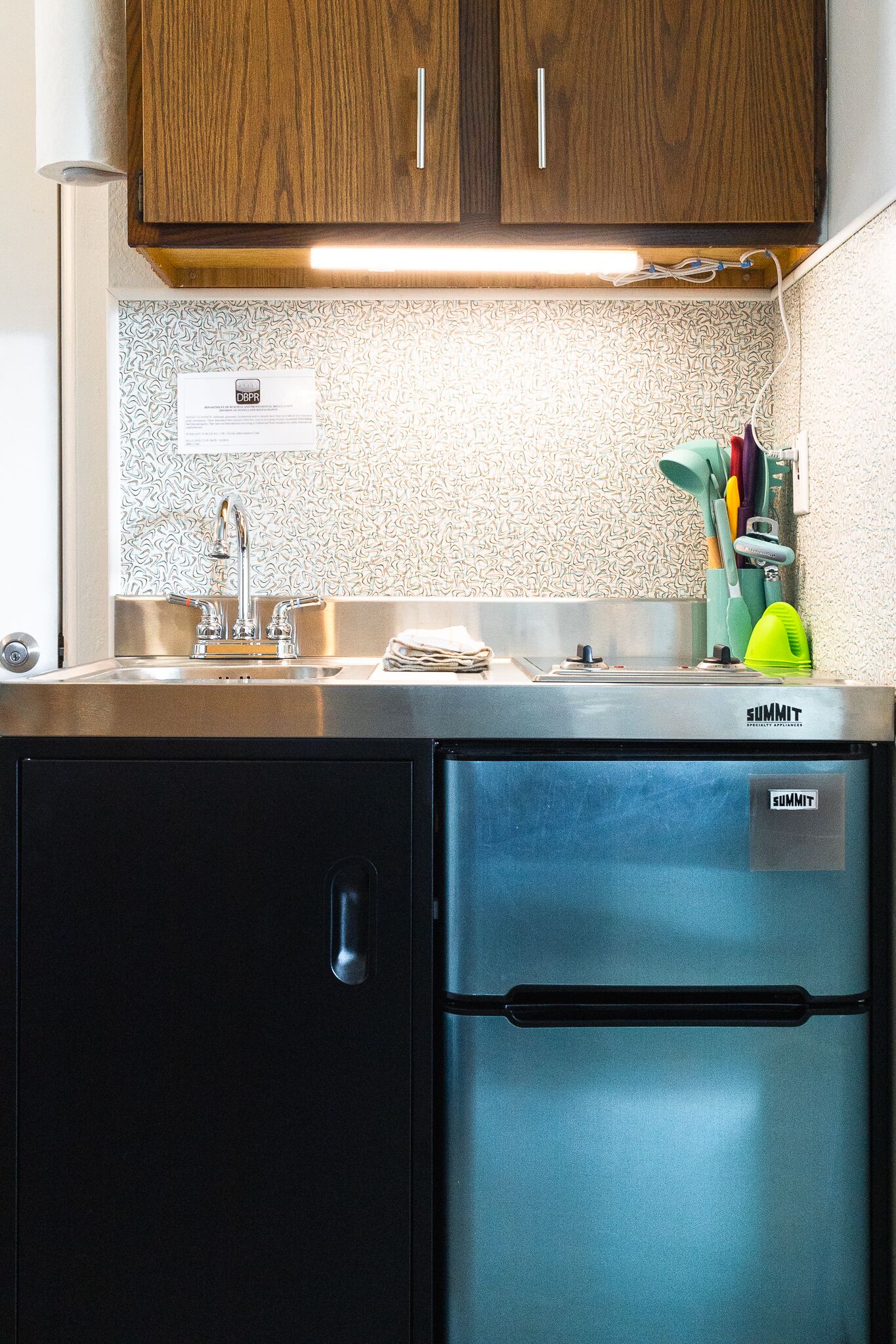Una cocina compacta con un refrigerador de acero inoxidable, un mueble de madera sobre el lavamanos y un salpicadero con diseño.