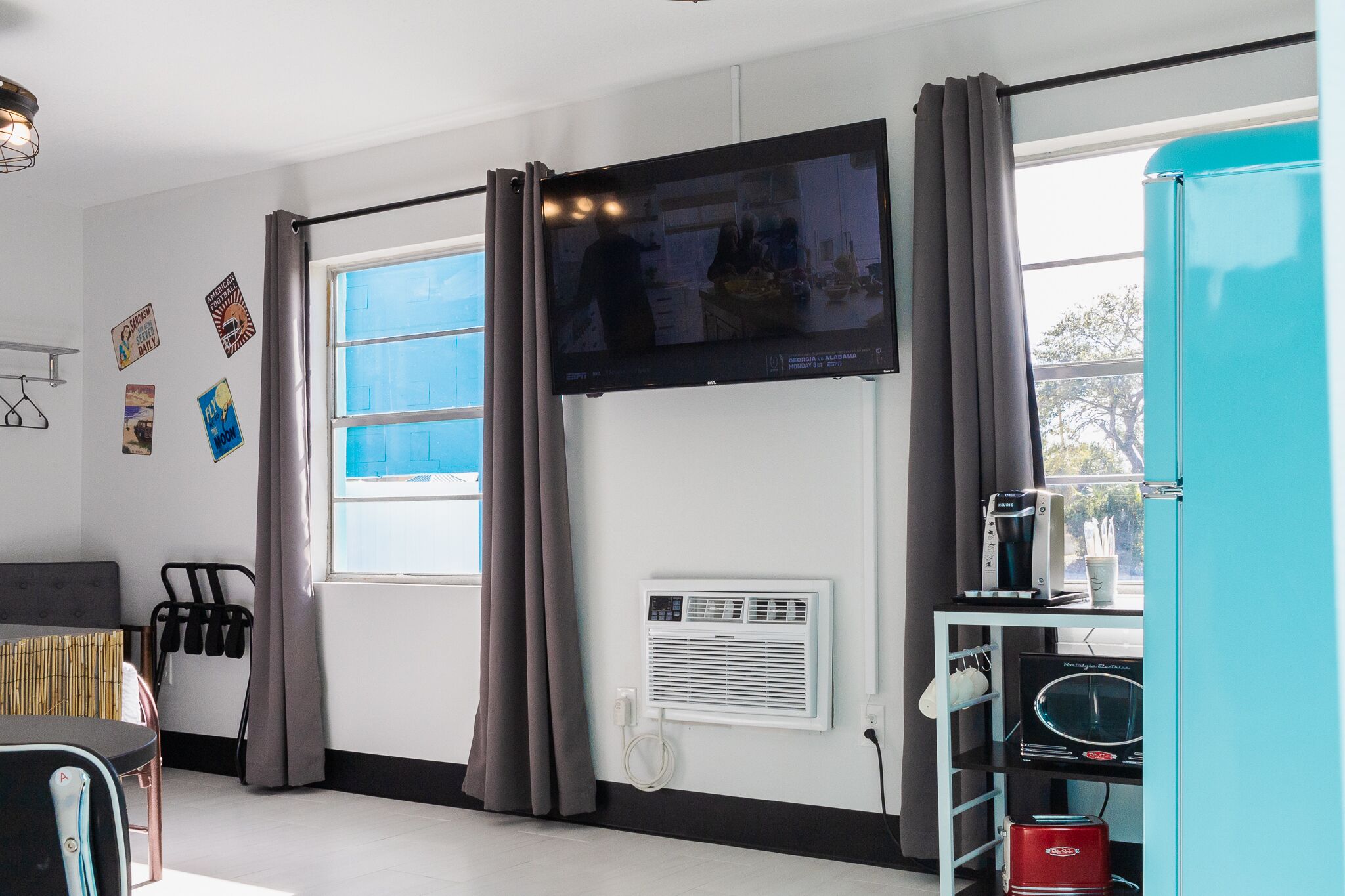A modern hotel room with a flat-screen TV mounted on the wall, a window with curtains, a wall-mounted air conditioner, and a small kitchenette area with a microwave and toaster.