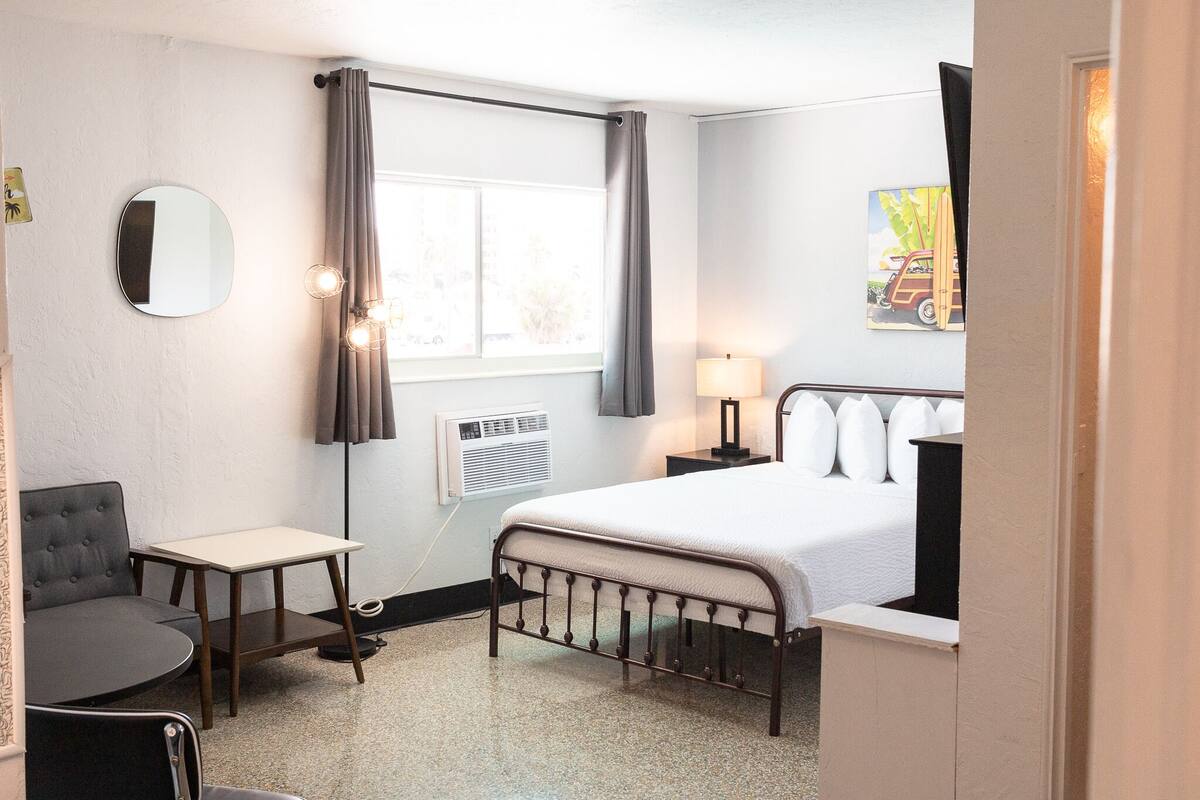 Hotellihuone, jossa on sänky, tuoli, pieni pöytä, peili, ikkuna verhoilla ja seinään kiinnitetty valaisin.