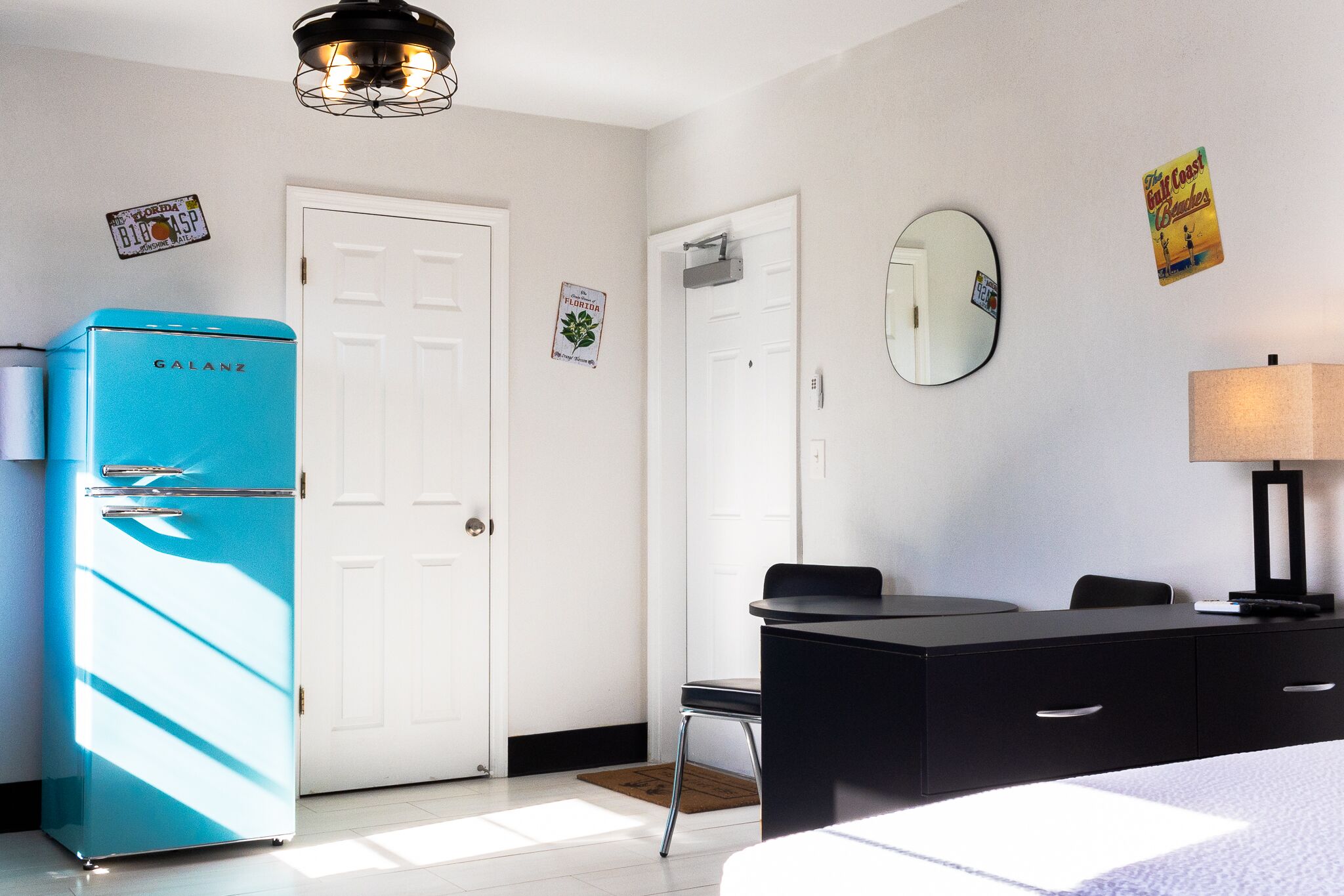 Une pièce avec un réfrigérateur bleu, un bureau avec une lampe, un miroir et une porte ornée d’une plaque d’immatriculation de la Floride.
