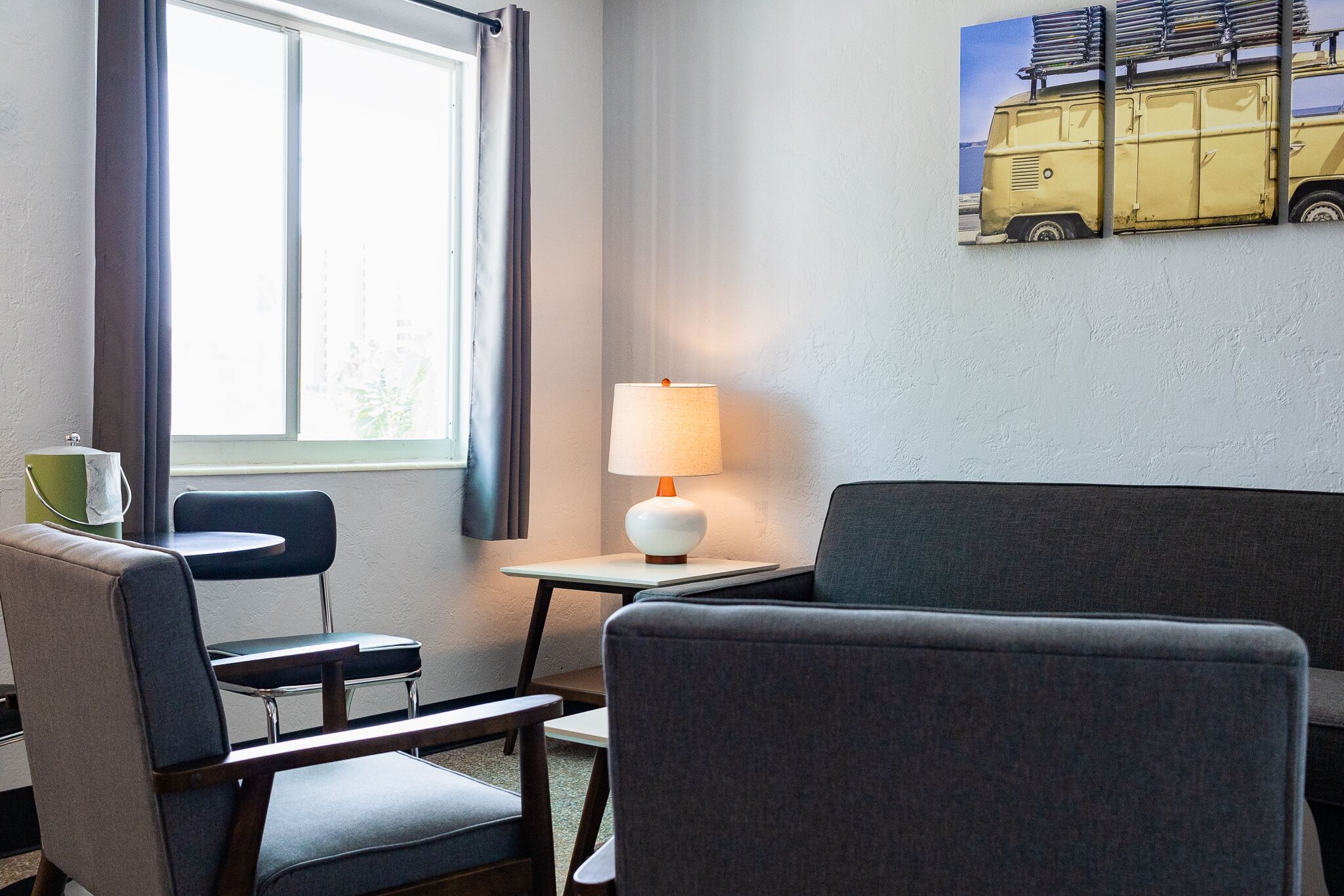 A hotel room with a sofa, two armchairs, a small table with a lamp, a window with curtains, and a wall-mounted picture of a van.