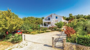 Una casa de dos pisos con balcón, rodeada de exuberante vegetación y un camino de entrada de grava.