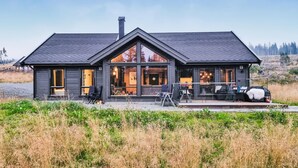 Moderni, kaksikerroksinen mökki, jossa on suuret ikkunat, katettu terassi ja savupiippu.