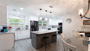 A modern kitchen with white cabinetry, a central island with bar stools, pendant lighting, and a dining area with a table and chairs. - Coastal Luxury - Walk to Beach - Heated Pool (Tarpon Springs)