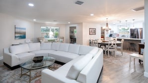 A modern living room with a white sectional sofa, a glass coffee table, and a dining area with a white table and chairs. - Coastal Luxury - Walk to Beach - Heated Pool (Tarpon Springs)
