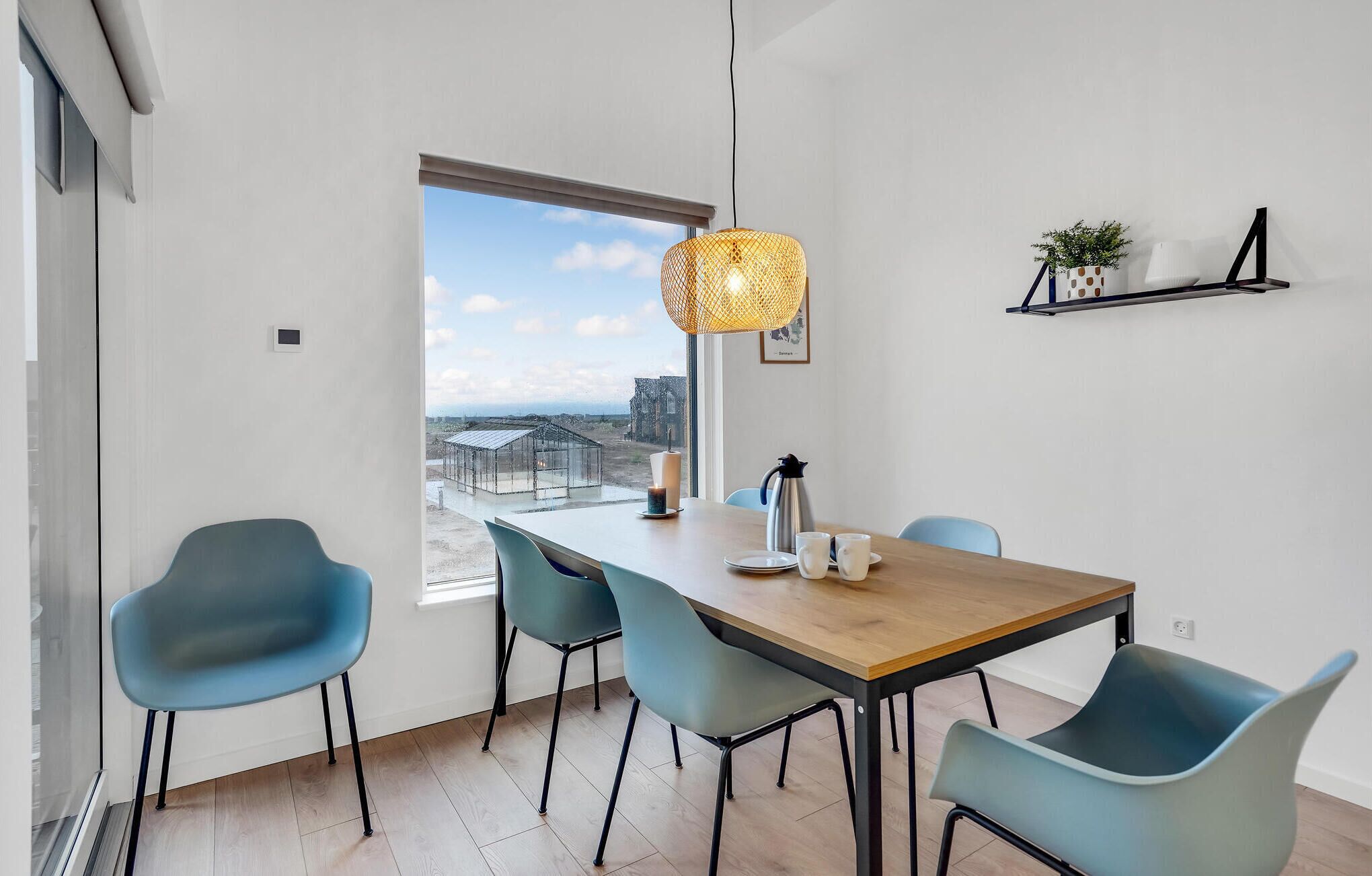 Een eethoek met een houten tafel, vier blauwe stoelen, een hanglamp, een kaars en uitzicht op een kas door een groot raam.