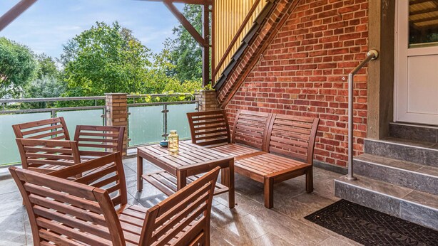 An outdoor patio with wooden furniture, a brick wall, and a view of greenery.