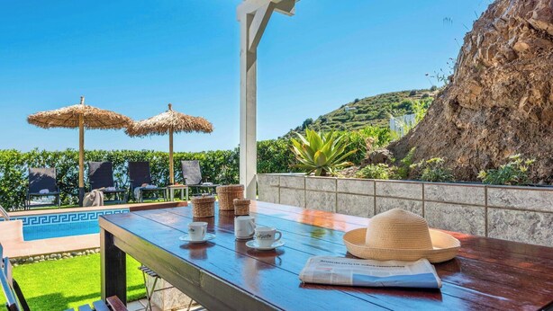 A wooden table with a newspaper, a straw hat, and a teapot set on a terrace with a pool and umbrellas.