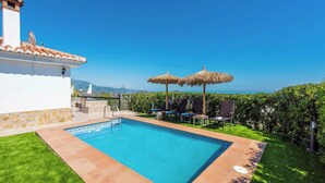 A swimming pool with a clear view of the sea, a white building with a tiled roof, and a grassy area with lounge chairs and umbrellas.