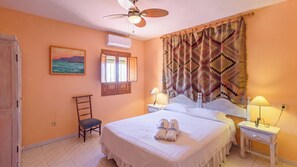 Habitación de hotel con cama, dos mesitas de noche, una silla, ventana con persianas, ventilador de techo y un cuadro en la pared.