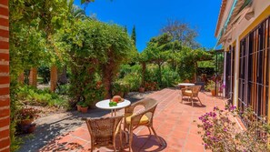 Un patio con mobiliario de mimbre y una mesa, rodeado de macetas con plantas y un muro de ladrillo.