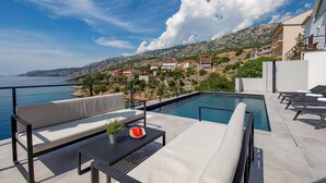 A poolside area with lounge chairs, a table with a watermelon and a plant, and a view of the sea and hillside buildings. - Stunning home in Prizna (Prizna)