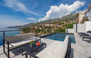 Ein Poolbereich mit Liegestühlen, einem Tisch mit Wassermelone und einer Pflanze, sowie Blick auf das Meer und Hügelhäuser.