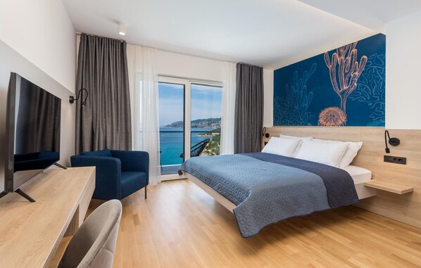 A hotel room with a bed, a desk, a chair, a TV, and a large window with a view of the sea.