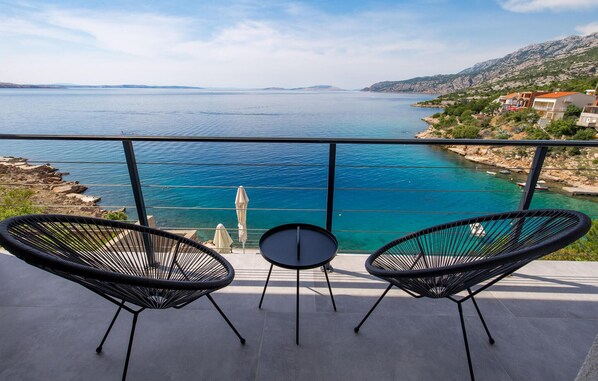 Ein Balkon mit zwei schwarzen Korbstühlen und einem kleinen runden Tisch, mit Blick auf eine Küstenlandschaft mit klarem, blauem Wasser und felsigem Gelände.