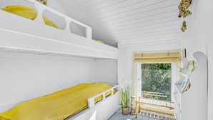 Habitació petita i senzilla, de tons blancs, amb lliteres, una finestra amb vistes a vegetació i una prestatgeria penjada a la paret.