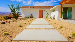 Una casa moderna con un camino de concreto que lleva a una puerta naranja, rodeada de plantas y rocas propias del desierto.