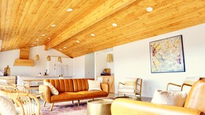 Una sala de estar moderna con techo de madera, un sofá, sillones, un comedor y una cocina al fondo.