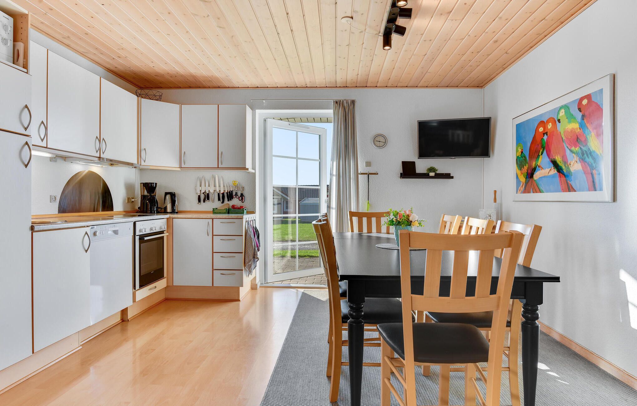 Une cuisine moderne avec des meubles blancs, un coin repas avec des chaises en bois, un téléviseur fixé au mur et un tableau coloré.
