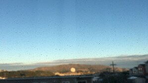 Blick durch ein Fenster mit Regentropfen, im Hintergrund ein Gebäude und ein klarer Himmel.