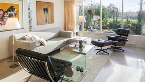 Un salon moderne avec un canapé, une table basse en verre, un fauteuil en cuir noir et une vue sur la verdure à l’extérieur.