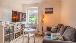 Ein gemütliches Wohnzimmer mit Fernseher, einem kleinen Tisch, einer Couch und einem Fenster mit Blick auf Grünflächen.
