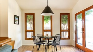 A dining area with a round table, four chairs, and a pendant light. There are three windows with wooden frames and a view of trees. A brown leather sofa is against the wall. - 5-Star Lux Modern Cabin in DT Fruita ❊ Dog Friendly❊ (Fruita)