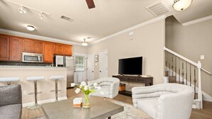 A spacious living room with a grey sofa, a coffee table, and a TV. There are three bar stools and a kitchen area with wooden cabinets and a microwave. - 8th #B xxxxxxxxxxxxx (Austin)