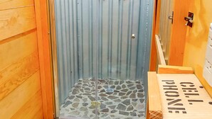 Um chuveiro pequeno e fechado, com chão de azulejos de pedra e um banco de madeira à direita.