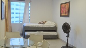 Une petite pièce comprenant une table en verre, deux chaises, un lit, un ventilateur sur pied et une fenêtre avec des rideaux.