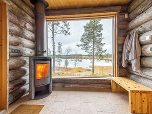 A log cabin interior with a wood-burning stove, a bench, and a large window offering a view of trees and a body of water. - Jussis chalet b by Interhome (Kuusamo)