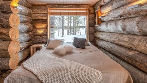 Interior de una cabaña de troncos con una cama, almohadas y una ventana que ofrece vistas a árboles y nieve.