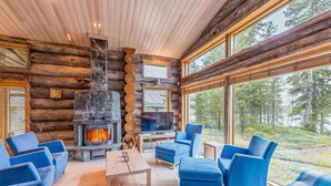 Interior de una cabaña de troncos con chimenea de piedra, muebles tapizados en azul y ventanas amplias con vista al bosque.