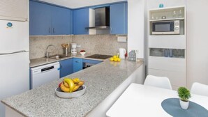 A modern kitchen with blue cabinets, a granite countertop, a built-in microwave, and a dining area with a white table and chairs. - Tramuntana by Interhome (L'Escala)