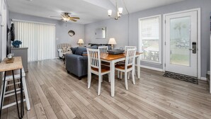 Un salon moderne avec un coin repas, agrémenté d’un ventilateur de plafond, d’un lustre et d’une grande fenêtre avec des stores.