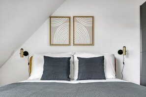 A modern bedroom with a sloped ceiling, a bed with white and gray bedding, two wall-mounted light fixtures, and two framed abstract wall art pieces.
