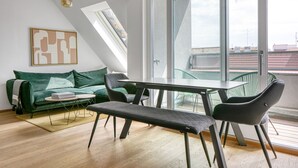 Un appartement moderne de style « loft », doté d’un coin repas, d’un canapé vert et d’une grande fenêtre offrant une vue sur la ville.