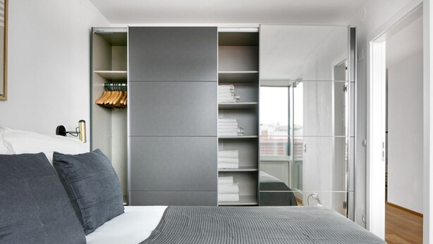 Une chambre moderne avec un lit, une literie grise, une armoire grise et une vue sur la ville par une grande fenêtre.