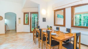 A dining area with a wooden table and chairs, a kitchen in the background, and large windows with a view of greenery. - Rei Sole (REI305) by Interhome (Costa Rei)