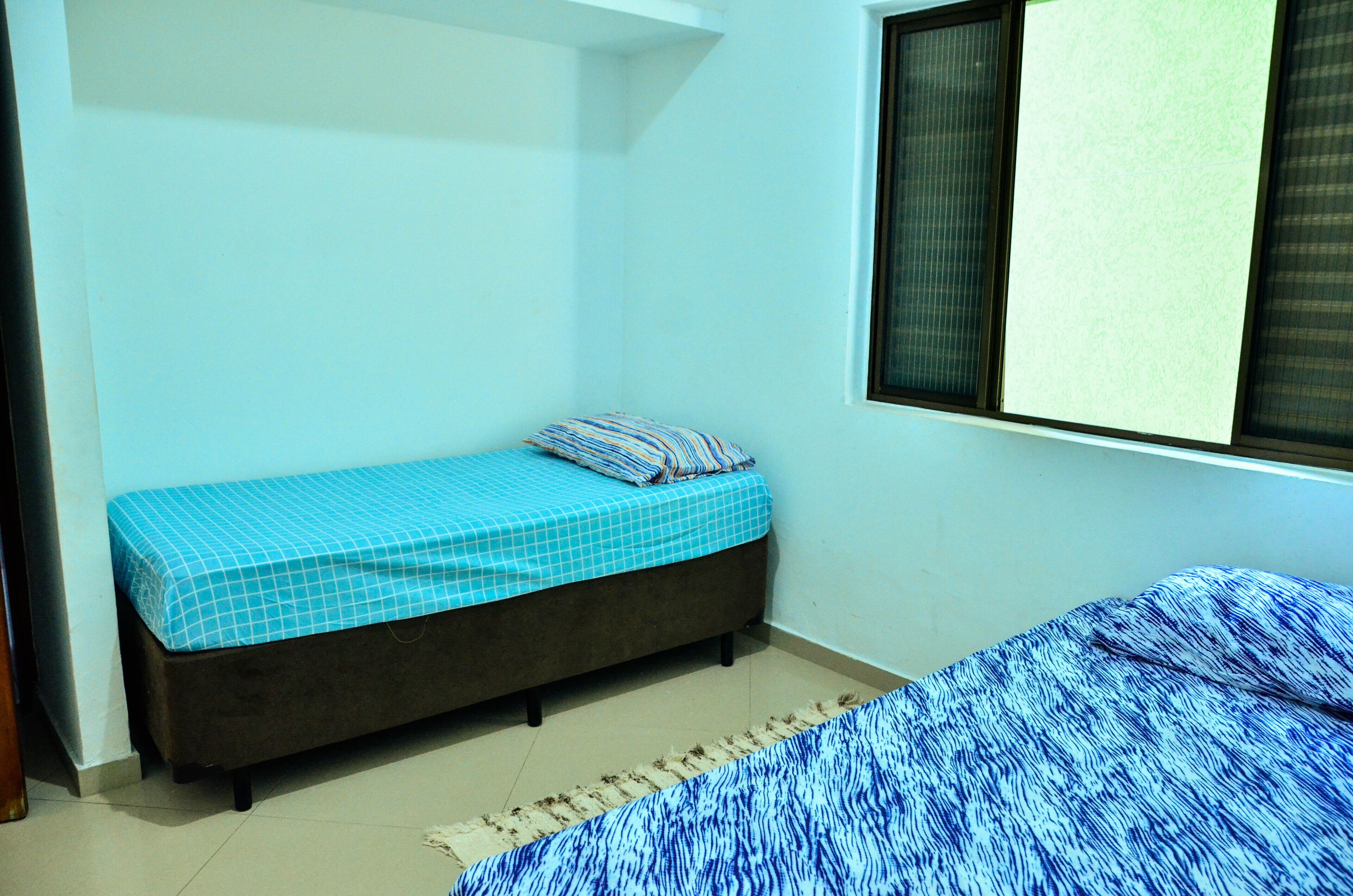 A small, single-bed room with a blue checkered bedspread and a matching pillow. The room has a window with blinds, a plain white wall, and a tiled floor.