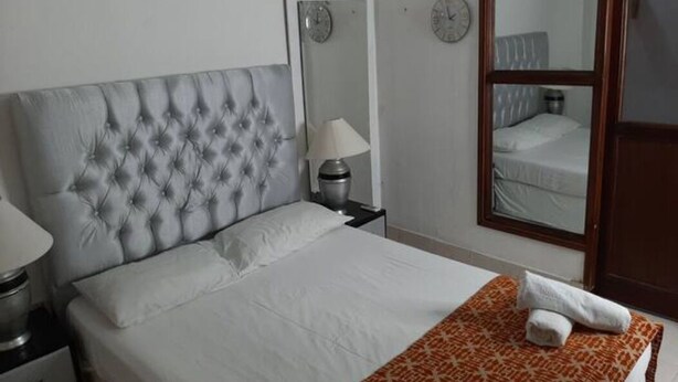 A hotel room with a tufted headboard, a bed with white linens, a bedside table with a lamp, a mirror, and a door. - 1G2-3 Apartamento en la ciudad Antigua (Cartagena)
