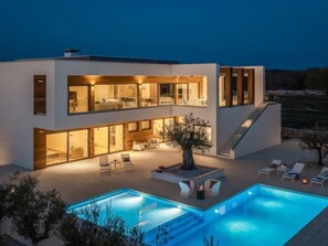 Une maison moderne à deux étages dotée d’une grande piscine extérieure, d’un escalier menant à l’étage supérieur et d’un patio bien éclairé.