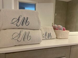 Une salle de bain avec un comptoir blanc, une serviette blanche ornée de lettres brodées et un panier contenant des objets décoratifs.