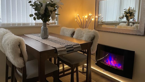 Un coin repas chaleureux avec une table en bois, des chaises blanches, un vase rempli de fleurs, une cheminée et un miroir au mur.
