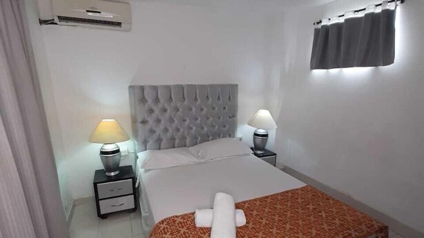 Habitación de hotel con cama, dos mesitas de noche con lámparas, ventana con cortinas y equipo de aire acondicionado en la pared.