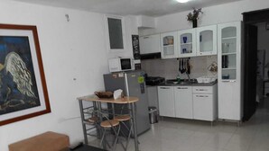 Una cocina compacta con armarios blancos, un microondas y un refrigerador. Hay un pequeño comedor con sillas y un sofá en la sala.