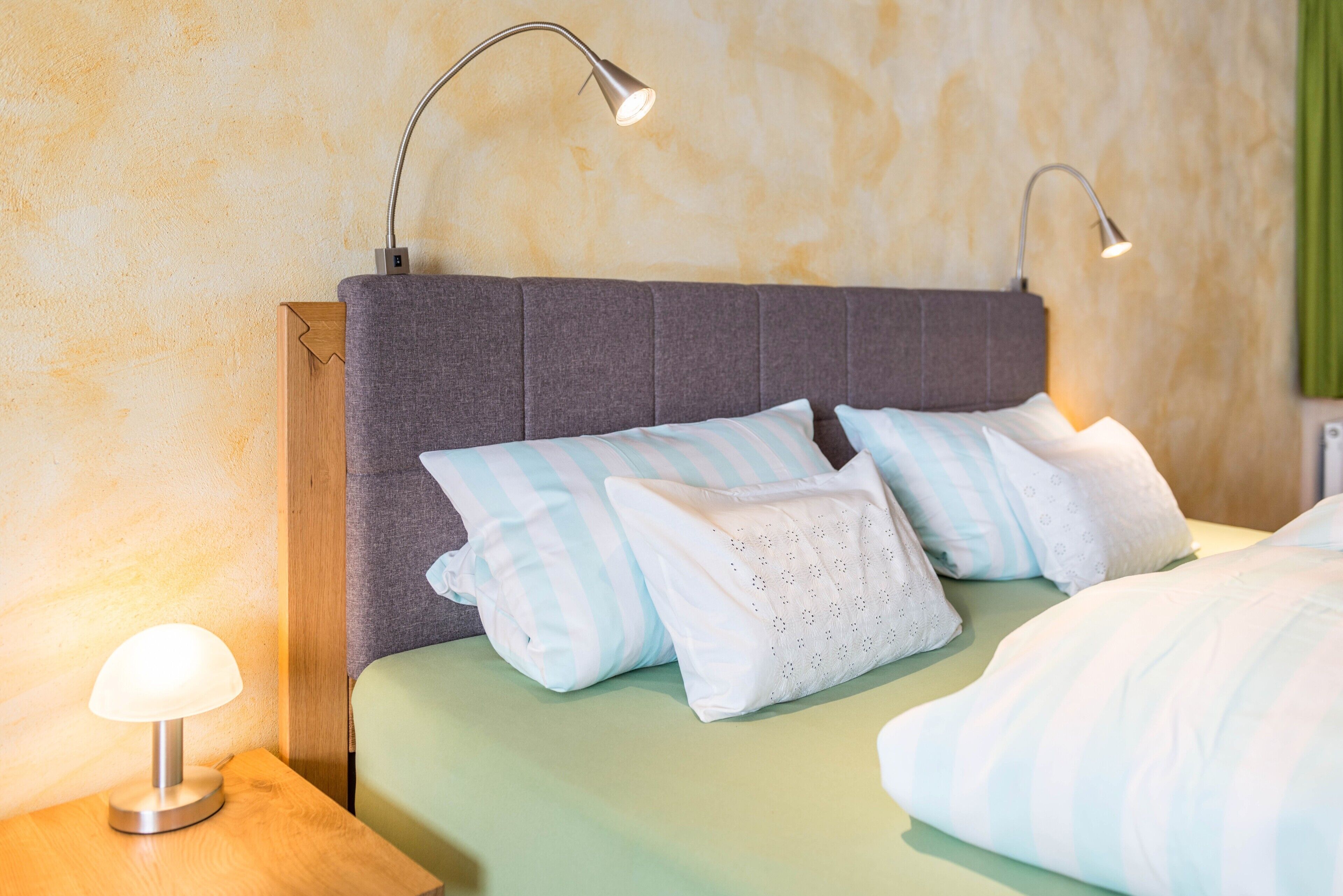 Een net opgemaakt bed met gestreepte kussens, een grijs hoofdbord en twee bedlampjes.