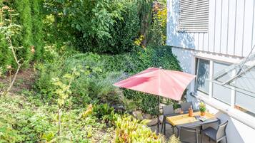 Een terras met een tafel en stoelen, omgeven door weelderige begroeiing en een rode parasol.