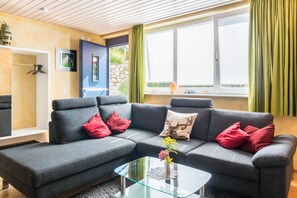 Un salon moderne avec un canapé d’angle, une table basse en verre et une vue sur l’extérieur grâce à de grandes fenêtres.